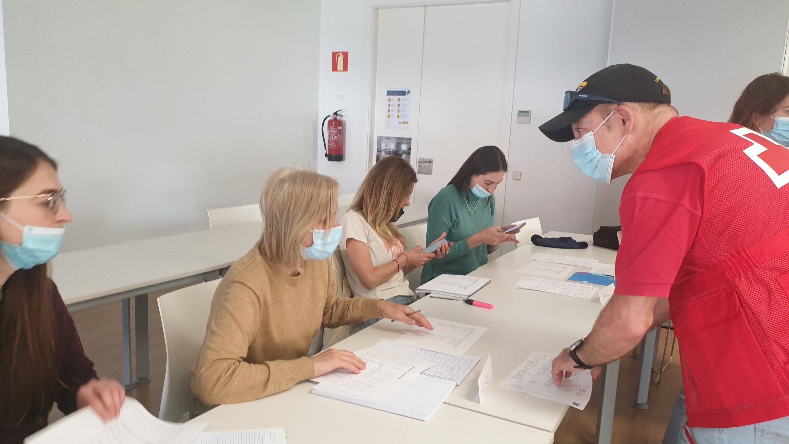 Miembro de Cruz Roja, de pie, hablando con alumnas sentadas en mesas.