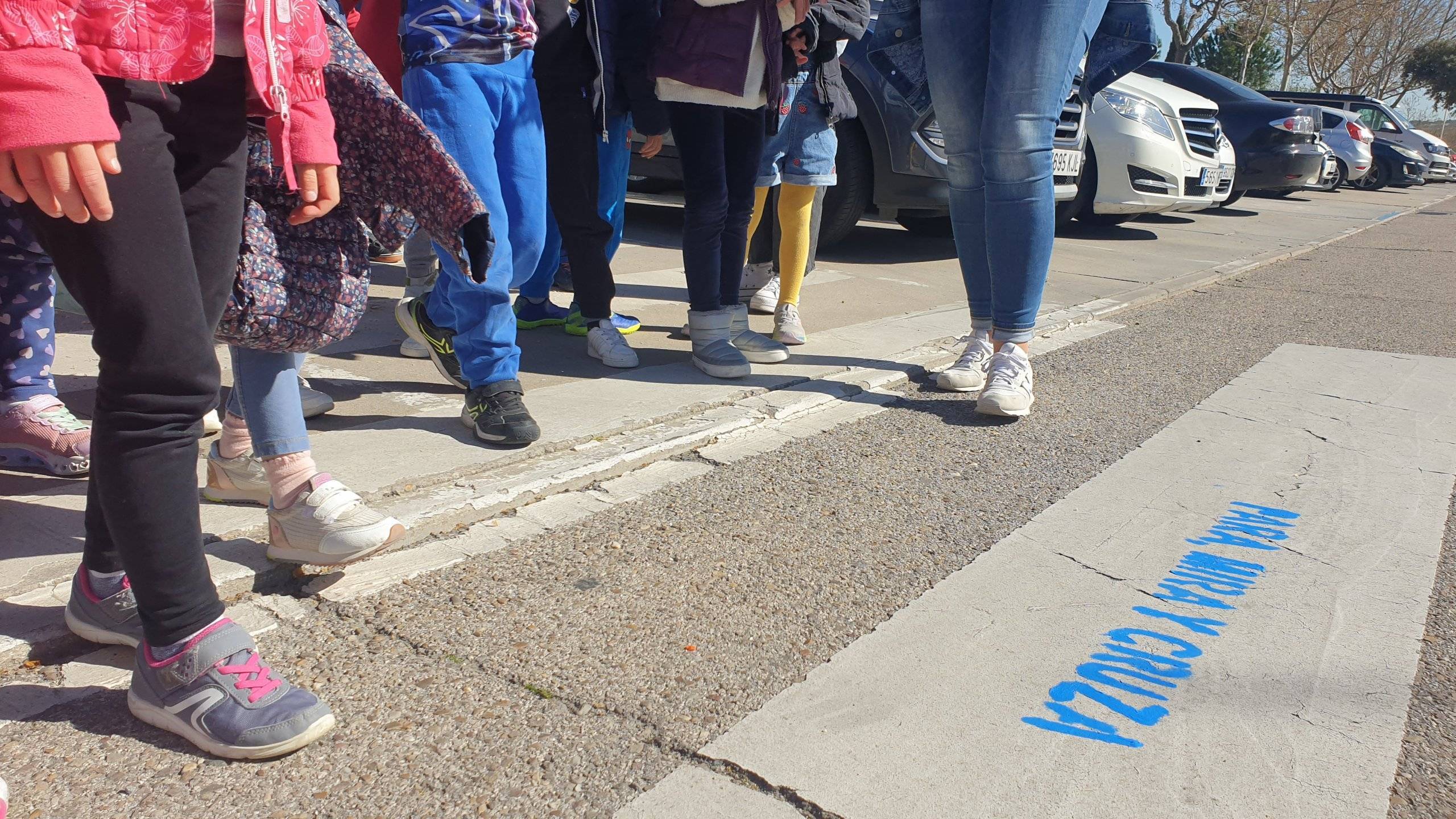 Niños esperando a cruzar en un paso de peatones en el que han pintado el mensaje: "Para, mira y cruza".