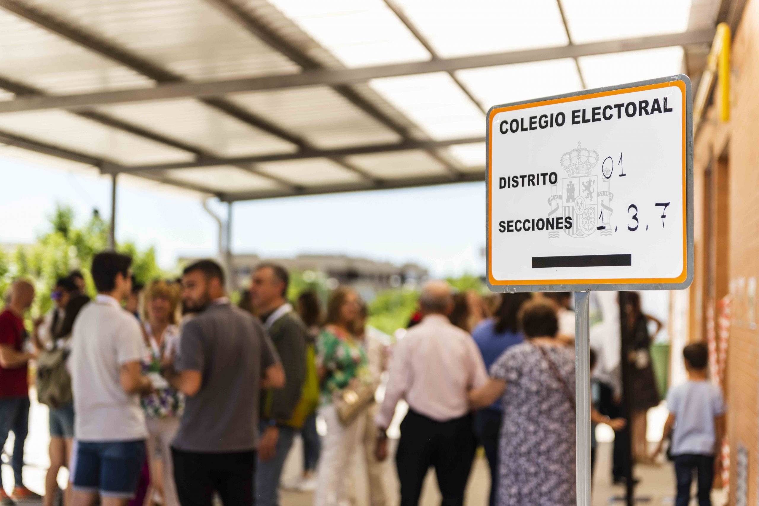 Imagen de cartel de colegio electoral en primer plano y gente al fondo.